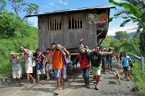 A group of men walk up a road towards the camera, carrying a wooden house on their shoulders.