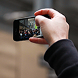 A thumbnail image of a hand holding a mobile phone in the air with a crown of people pictured on the screen.
