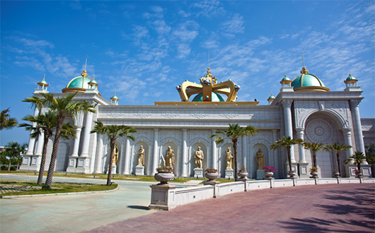 Cream-coloured casino building with palm trees and gold and turquoise painted domes and crown on the roof and gold-coloured statues at the front.