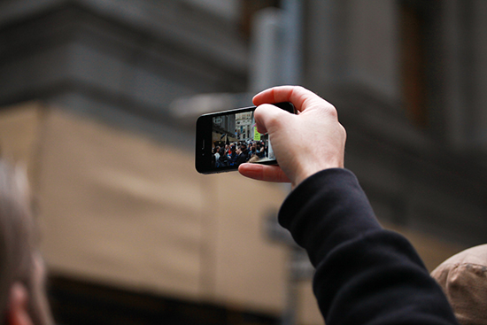 A man's hand holding a mobile phone up in the air. On the screen of the phone is a crowd of people in the street.