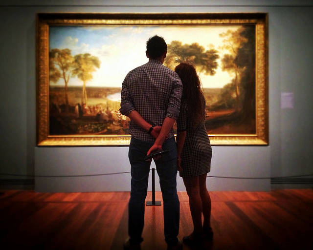 A man and a woman look at a landscape painting, with their backs to the camera.
