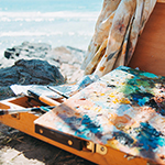An abstract painting on canvas and a wooden box of art supplies sits on the rocks near the ocean. 