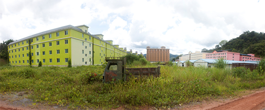 An abandoned truck and overgrown grass surrounds large buildings. 