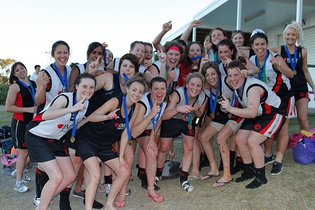 With medals around their necks, members of a women's AFL team smile for the camera, making 'Number 1' signs with their hands.