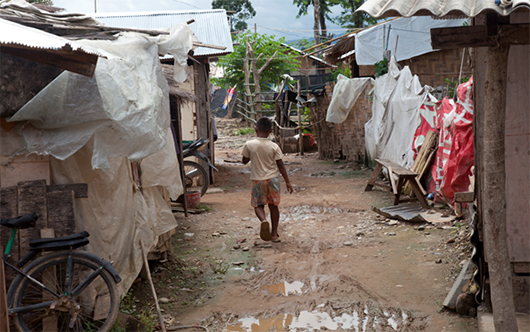 A young boy walks along a muddy path between roughly constructed shelters with large sheets of plastic hanging from them.
