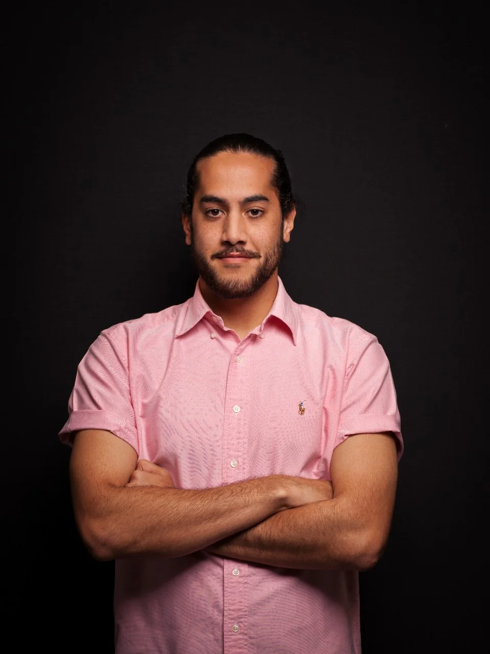 Male student wearing a pink shirt with arms crossed smiling at the camera