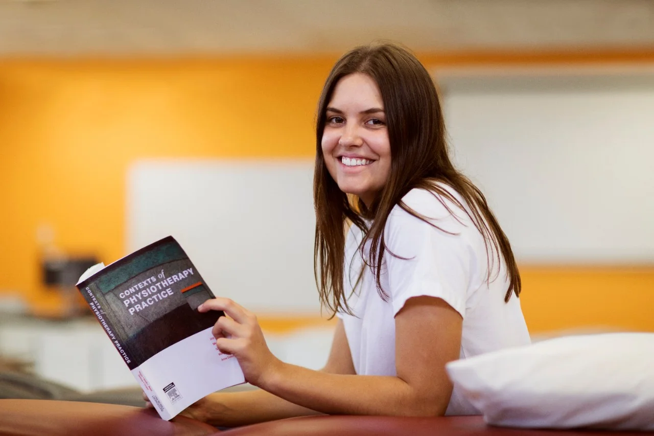 Female student holding a text book and smiling at the camera